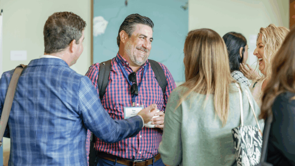 A group of people stand in a circle indoors, smiling and conversing at a professional event. One man in the center wears a checkered shirt and name badge, holding a coffee cup.