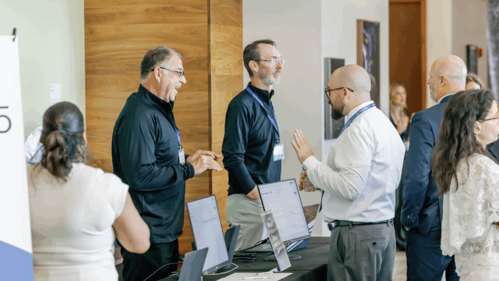 People engage in conversation around a booth with computer monitors at a healthcare event, with several attendees wearing conference badges and lanyards.